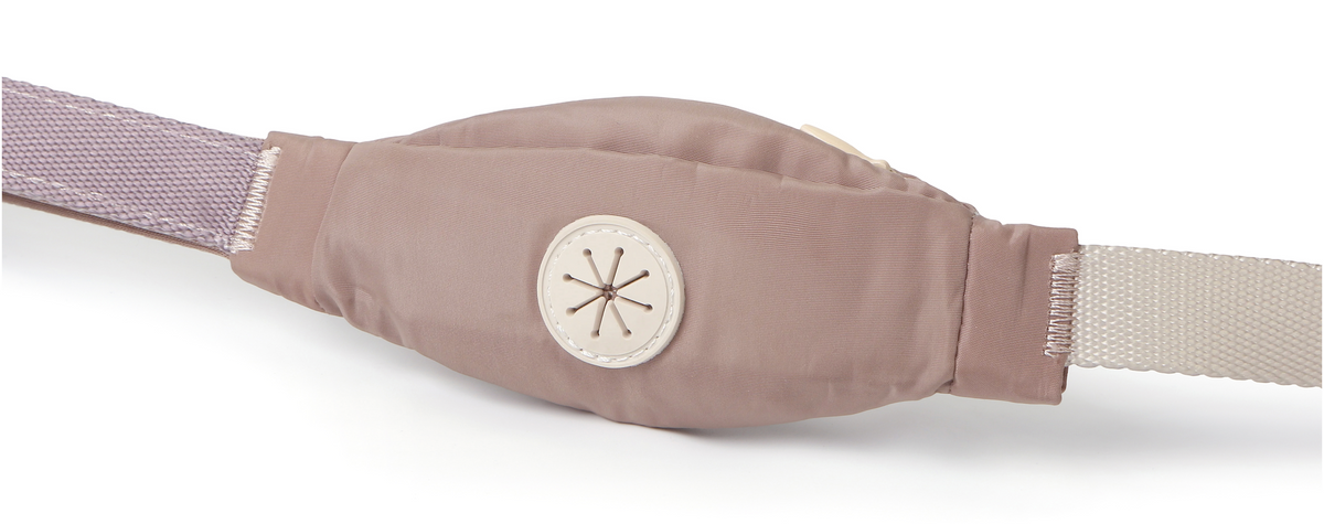 A close-up of a beige fabric waist wrap or belt with a central circular button closure and a light purple woven strap on one side and a beige strap on the other, shown against a plain white background.